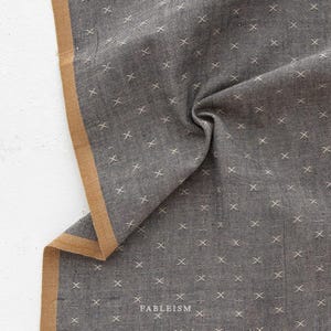 May include: A close-up of a gray fabric with a white cross pattern. The fabric has a tan border and the text "FABLEISM" is printed in white on the fabric.