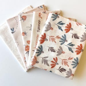 May include: A set of five folded fabric squares in cream, featuring various printed patterns including blue dragonflies, orange strawberries, and floral motifs in shades of rust and blue, with the text Summer joy printed on one piece.