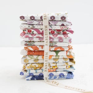 May include: A vertical stack of folded fabric squares featuring various floral and botanical patterns in shades of purple, red, orange, yellow, and blue. A cream-colored ribbon printed with the text FABLEISM wraps around the fabric stack.