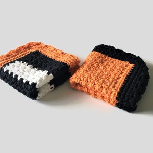 May include: Two crocheted dishcloths in orange, black, and white. The dishcloths are made with a textured stitch.