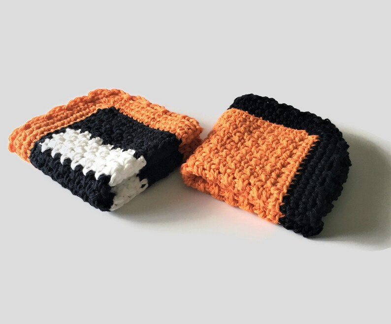 May include: Two crocheted dishcloths in orange, black, and white. The dishcloths are made with a textured stitch.