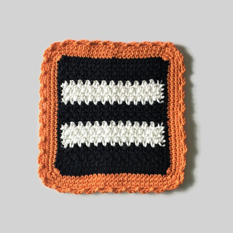 May include: A crocheted dishcloth with an orange border and two white stripes on a black background.