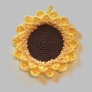 May include: A crocheted sunflower pot holder with yellow petals and a brown centre. The centre is surrounded by a ring of orange and yellow yarn.