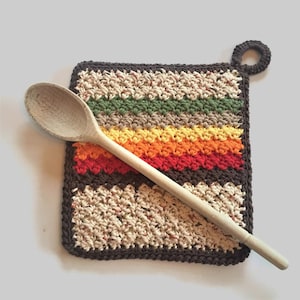 May include: A brown crocheted pot holder with a hanging loop. The pot holder has stripes of green, beige, yellow, orange, and red yarn. A wooden spoon is lying on top of the pot holder.