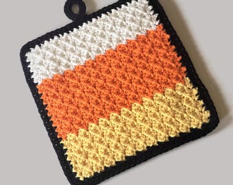 May include: A crocheted pot holder in the shape of a candy corn. The pot holder is made with black, white, orange, and yellow yarn. It has a loop handle for hanging.