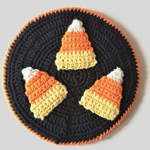 May include: A black crocheted circle with an orange border features three crocheted candy corn designs in orange, yellow, and white.