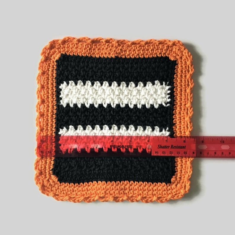 May include: A black and white striped crocheted dishcloth with an orange border. The dishcloth is approximately 6 inches wide and 6 inches long.
