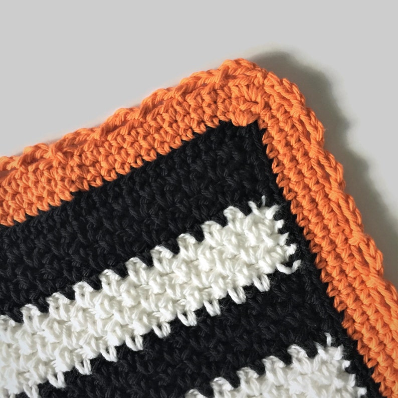 May include: A crocheted dishcloth with black, white, and orange stripes. The dishcloth has a crocheted border in orange.