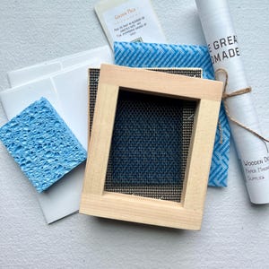 K&ouml;nnte beinhalten: Ein Holzrahmen f&uuml;r die handgemachte Papierherstellung, mit einem blauen Schwamm, einer Rolle wei&szlig;en Papiers und einem kleinen P&auml;ckchen goldenem Glimmer. Die Papierherstellungsmaterialien befinden sich auf einer wei&szlig;en Oberfl&auml;che.