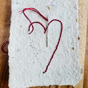 K&ouml;nnte beinhalten: Ein wei&szlig;es Papierjournal mit einem roten Herz, das auf die Vorderseite gestickt ist. Das Herz ist mit rotem Garn gestickt und eine Nadel steckt noch im Papier.