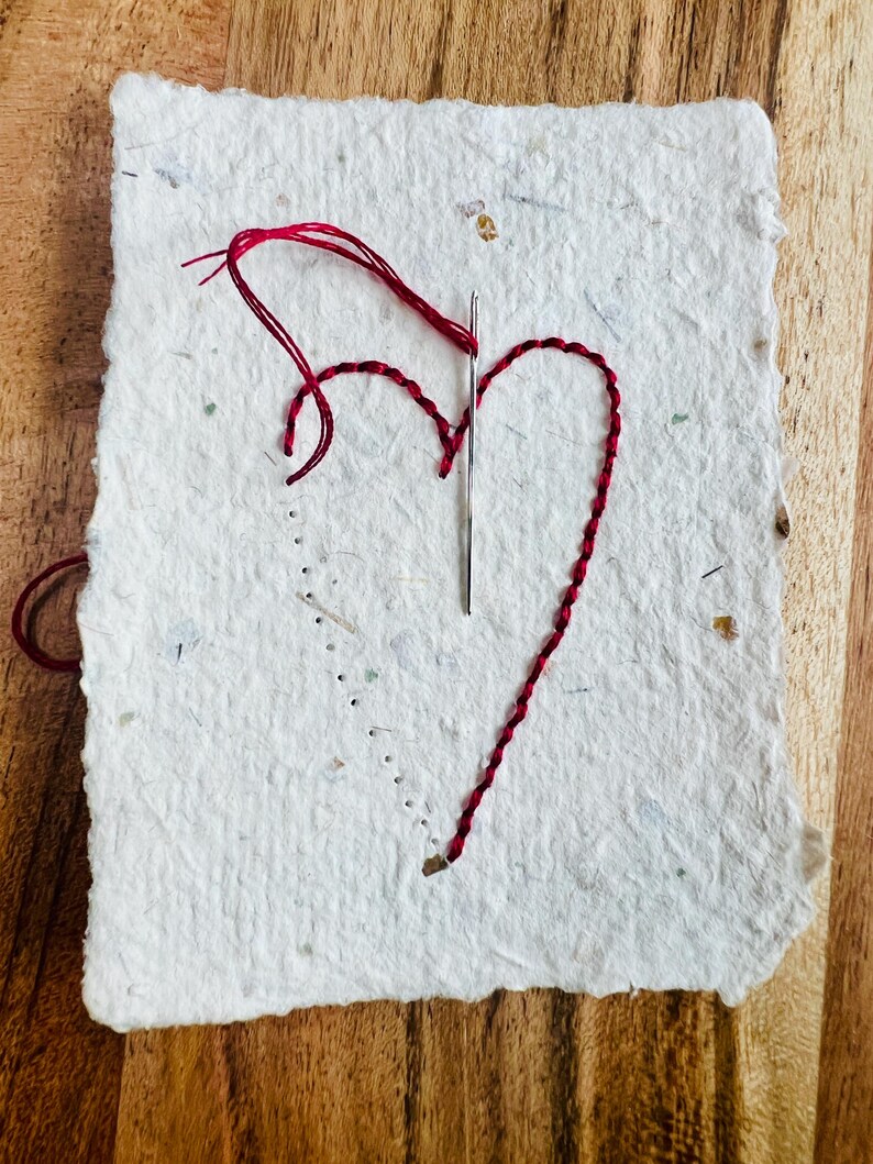 K&ouml;nnte beinhalten: Ein wei&szlig;es Papierjournal mit einem roten Herz, das auf die Vorderseite gestickt ist. Das Herz ist mit rotem Garn gestickt und eine Nadel steckt noch im Papier.