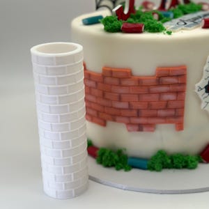 May include: A white, cylindrical cake decoration resembling a brick wall stands in front of a decorated cake. The cake features a brick wall design, green frosting, and a Spider-Man theme.