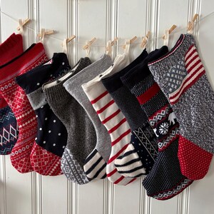 May include: A collection of nine knitted Christmas stockings in various colors and patterns, including red, navy, gray, and white. The stockings are hung on a string with wooden clothespins against a white paneled wall. Two woven baskets are above.