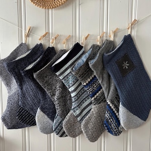 May include: A collection of eight knitted Christmas stockings in various shades of blue, gray, and white, hanging from a string with wooden clothespins. The stockings feature different patterns and textures, including cable knit and geometric designs. Two woven wall decorations are above the stockings.