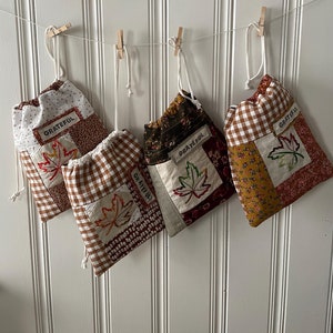 May include: Four fabric drawstring bags with a patchwork design featuring brown, white, and orange gingham patterns, floral prints, and a single embroidered maple leaf on each bag. The bags are hung on a white string with clothespins and each bag has a label that reads "Grateful".