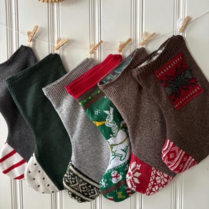 May include: A collection of Christmas stockings in various colors and patterns, including gray, green, and brown, are hung with wooden clothespins. Some stockings feature festive designs like snowflakes and a snowman.