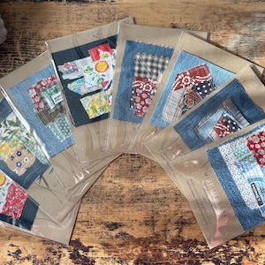 May include: A collection of handmade fabric art pieces, each featuring a unique patchwork design on denim. The pieces are packaged in clear plastic sleeves with kraft paper backing, arranged in a fan shape on a wooden surface. The patchwork includes various colors and patterns.