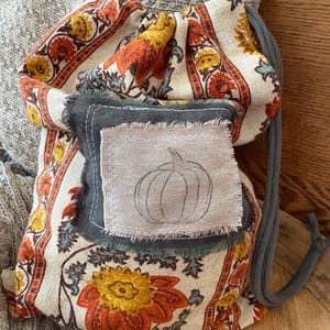 May include: A drawstring bag with a floral pattern in orange, yellow, and red. A gray patch with a pumpkin sketch is sewn on the front. The bag has a gray drawstring and is resting on a wooden surface.