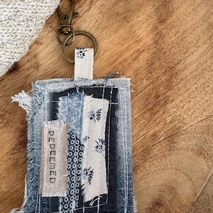 May include: A handmade denim keychain with a brass clasp. The rectangular tag features layered fabric patches in shades of blue and white, with the word "REDEEMED" visible. The keychain is on a wooden surface.