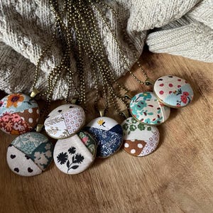 May include: A collection of eight necklaces with round fabric pendants. The pendants are made of different patterned fabrics in shades of blue, green, red, white, and brown. The necklaces are made of gold-toned chains.