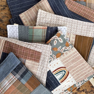 May include: A collection of handmade fabric coasters in various patterns and colors. The coasters feature patchwork designs with stripes, plaid, and floral motifs. Colors include blue, brown, cream, and green. The coasters are arranged on a wooden surface.