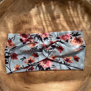 May include: A light blue headband with a floral pattern in shades of pink and brown. The headband is twisted and has a soft, stretchy fabric.