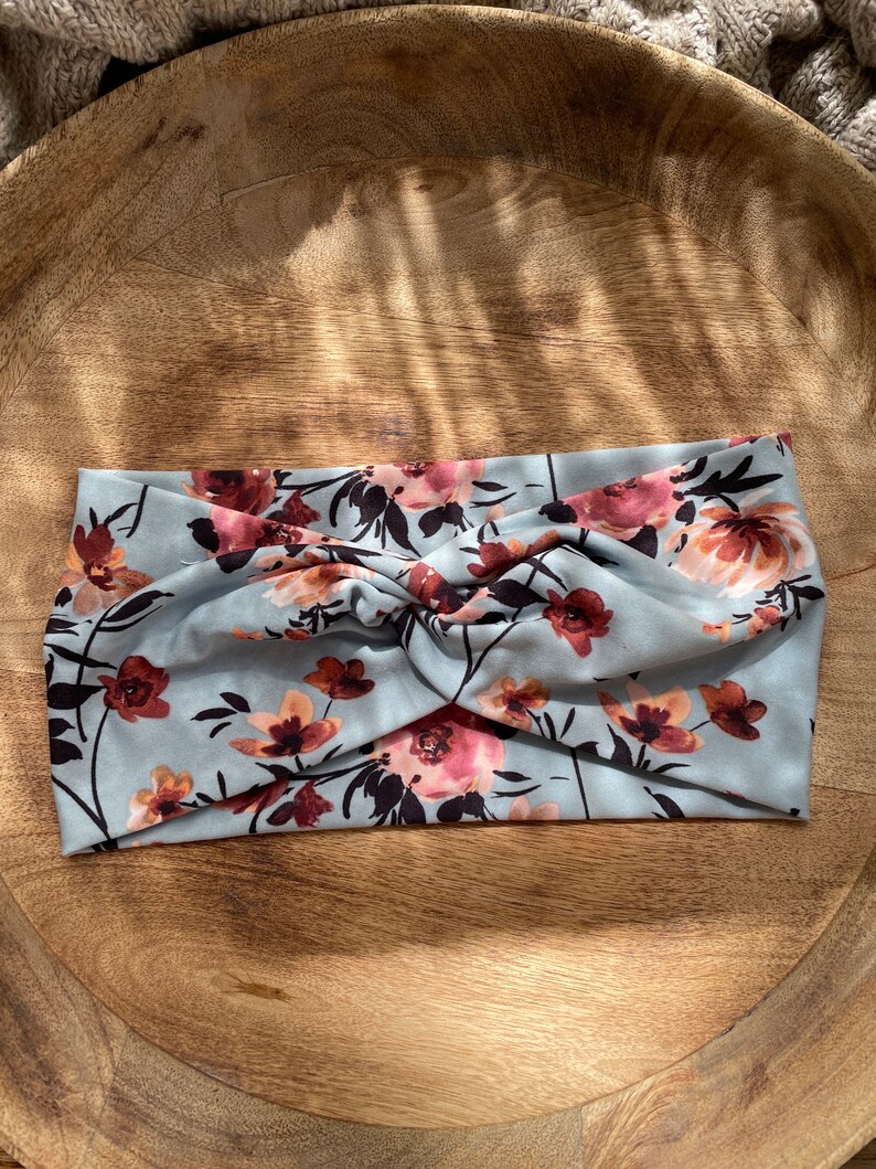 May include: A light blue headband with a floral pattern in shades of pink and brown. The headband is twisted and has a soft, stretchy fabric.