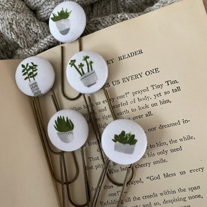 May include: Set of five fabric-covered paper clips featuring small, hand-drawn potted plants in shades of green and gray. The clips are attached to a vintage book page, with a gray knitted blanket in the background. A charming, botanical-themed stationery item.