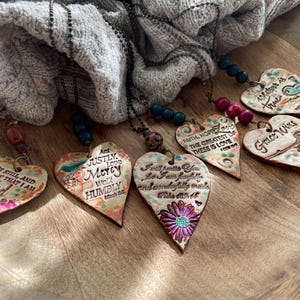 May include: Five heart-shaped ceramic pendants with inspirational quotes and floral designs. The pendants are on brown chains with various colored beads.