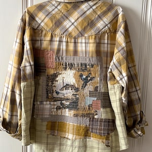May include: A long-sleeve button-down shirt in shades of mustard yellow, white, and brown plaid. The back features a large, rectangular fabric patch with a printed landscape scene of a house and birds, surrounded by smaller, layered plaid fabric scraps.