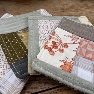 May include: A set of three square fabric coasters featuring patchwork designs in shades of olive green, grey, white, and burnt orange. The coasters are constructed with visible straight-line quilting stitches and finished with solid grey fabric borders.