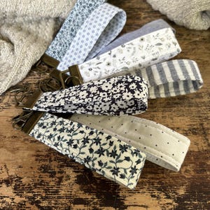 May include: A collection of fabric wrist keychains with various patterns, including floral, polka dot, and striped designs. The keychains feature brass-colored hardware and are arranged on a wooden surface. The color palette includes blue, white, and black.