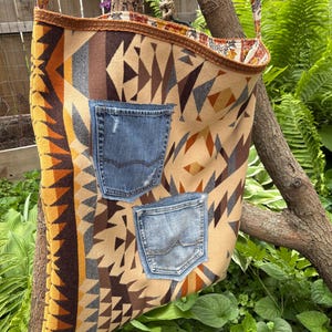 May include: A beige and brown patterned shoulder bag with two denim pockets. The bag has a floral patterned strap and trim. The bag is hanging from a tree branch in a garden setting.