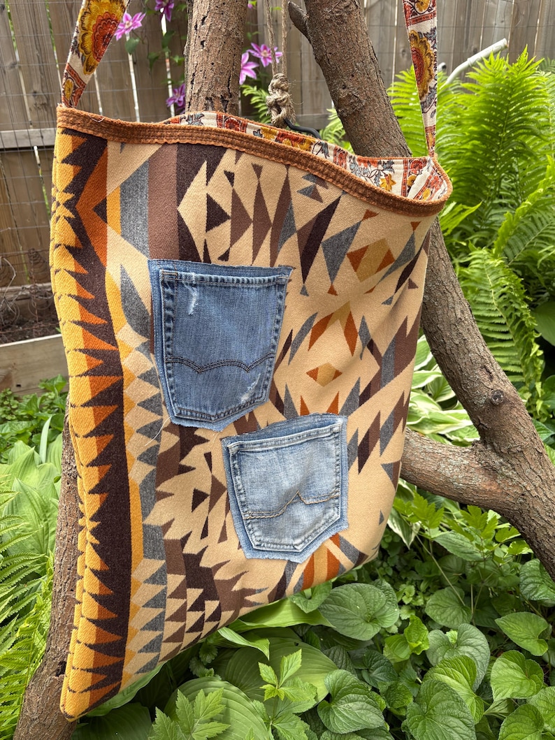 May include: A beige and brown patterned shoulder bag with two denim pockets. The bag has a floral patterned strap and trim. The bag is hanging from a tree branch in a garden setting.