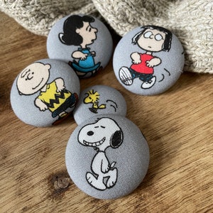 May include: Five round fabric buttons featuring characters from the Peanuts comic strip. The buttons are gray with white outlines and feature Charlie Brown, Snoopy, Lucy, Linus, and Woodstock.