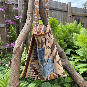 May include: A triangular tote bag with a patterned design in shades of brown, tan, and orange, featuring two denim pockets. The bag has a strap with a floral pattern and is hanging from a tree branch. The bag's dimensions are approximately 15 inches wide and 20 inches tall.