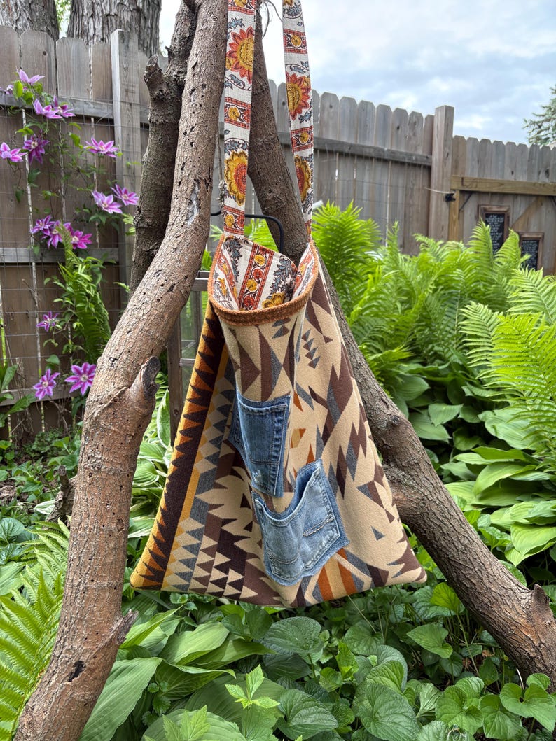 May include: A triangular tote bag with a patterned design in shades of brown, tan, and orange, featuring two denim pockets. The bag has a strap with a floral pattern and is hanging from a tree branch. The bag's dimensions are approximately 15 inches wide and 20 inches tall.