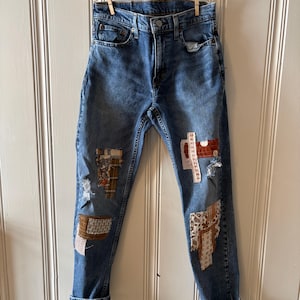 May include: Blue denim jeans with decorative patches in various patterns and colors. The jeans are hanging on a clothesline with wooden clothespins. The cuffs are rolled up, and the jeans have a classic five-pocket design.