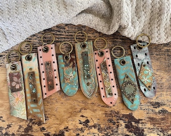 May include: A collection of handmade keychains with brass rings. Each keychain features a unique design with painted details in shades of pink, teal, and brown. The keychains are made of wood and have a rustic, artistic aesthetic.