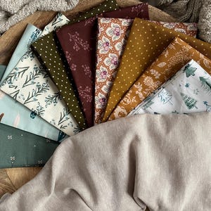 May include: A collection of 10 fabric squares with various patterns and colors, including green, brown, white, and gold. The fabrics are arranged in a fan shape on a wooden surface.