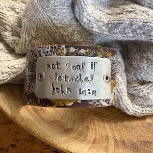May include: A wide leather cuff bracelet with a brown, yellow, and white patterned design. A silver metal plate is riveted to the front, with the words "not good if detached John 15:4" stamped on it.