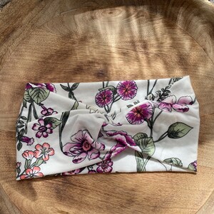 May include: A white headband with a floral pattern featuring pink and purple flowers. The headband is made of a soft, stretchy fabric and is designed to be worn as a hair accessory.