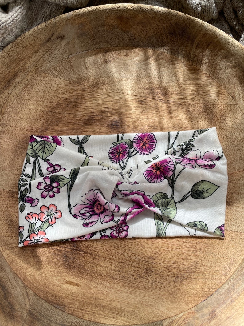 May include: A white headband with a floral pattern featuring pink and purple flowers. The headband is made of a soft, stretchy fabric and is designed to be worn as a hair accessory.