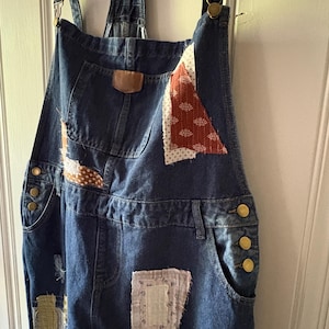 May include: Blue denim overalls with patchwork details. The overalls feature various fabric patches in shades of red, white, brown, and green. Brass-colored buttons and adjustable straps complete the design. The overalls are hanging against a white door.