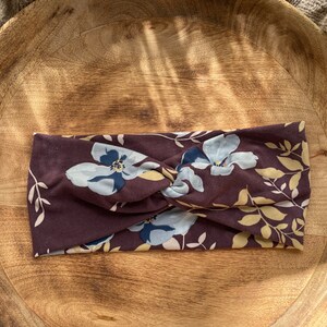 May include: A brown headband with a floral pattern featuring blue and yellow flowers. The headband is made of a soft, stretchy fabric and is designed to be worn as a hair accessory.