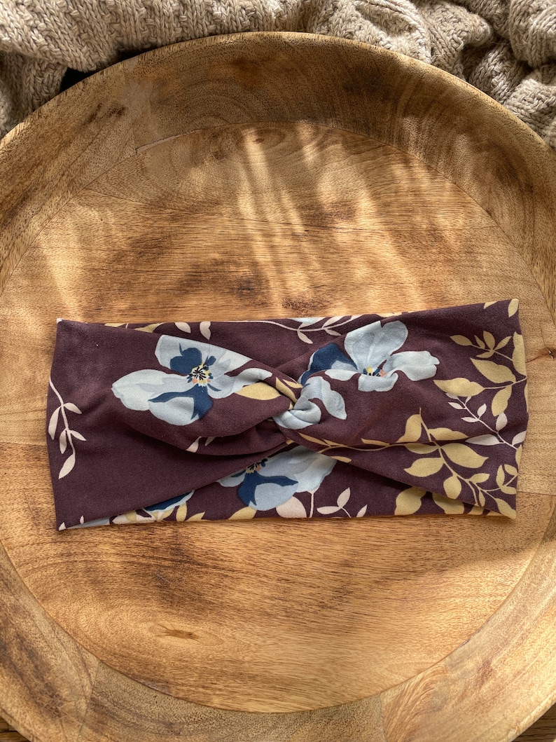 May include: A brown headband with a floral pattern featuring blue and yellow flowers. The headband is made of a soft, stretchy fabric and is designed to be worn as a hair accessory.