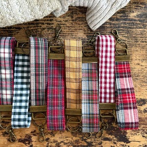 May include: A collection of fabric wrist keychains with various plaid patterns, including red, black, and yellow. Each keychain has a bronze-colored clasp and ring for attaching keys.