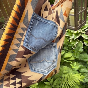 May include: A handmade tote bag with a geometric pattern in shades of brown, tan, and orange. Two denim pockets are sewn onto the front. The bag is hanging in a garden setting with lush green foliage.