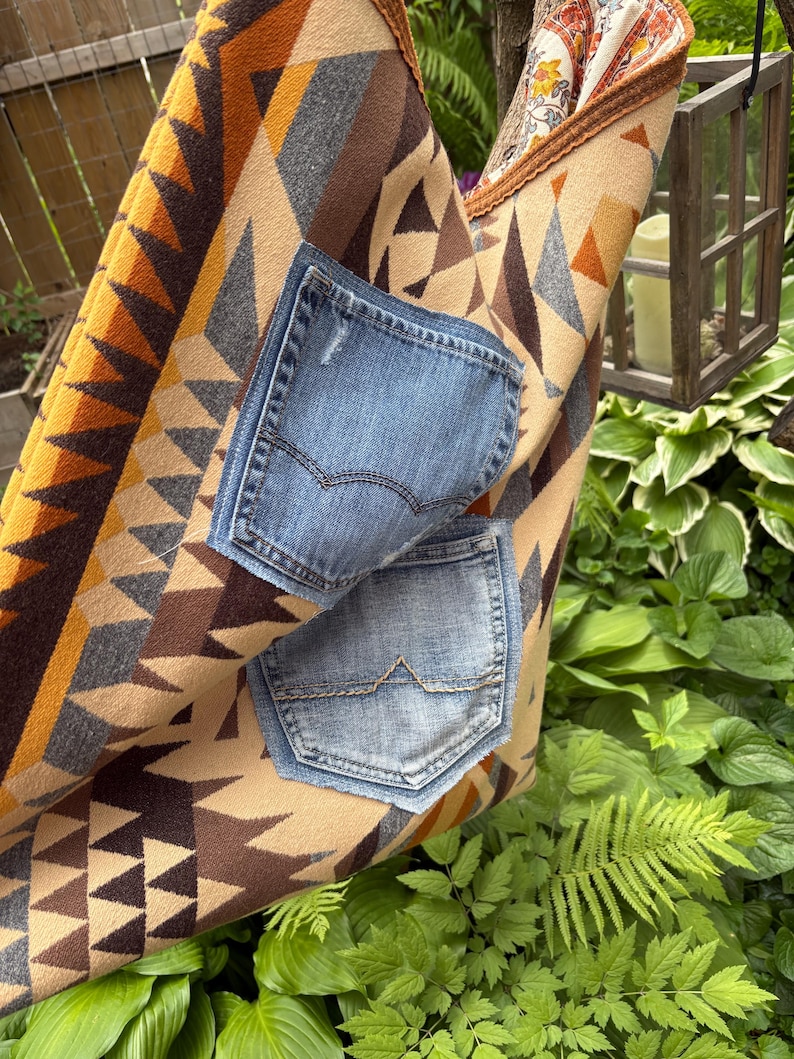 May include: A handmade tote bag with a geometric pattern in shades of brown, tan, and orange. Two denim pockets are sewn onto the front. The bag is hanging in a garden setting with lush green foliage.