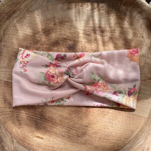 May include: A pink floral headband with a twisted knot design. The headband features a variety of pink, yellow, and green floral patterns.